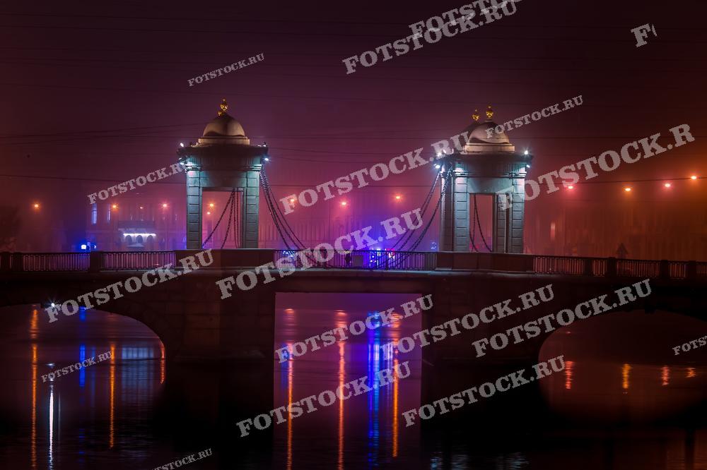 Фотография показывает мост Ломоносова в Санкт-Петербурге, окутанный густым туманом. Мягкий свет фонарей создаёт загадочную и таинственную атмосферу, отражаясь в водах Фонтанки. Это изображение подходит для использования в материалах, посвященных городским пейзажам и ночной жизни Санкт-Петербурга.